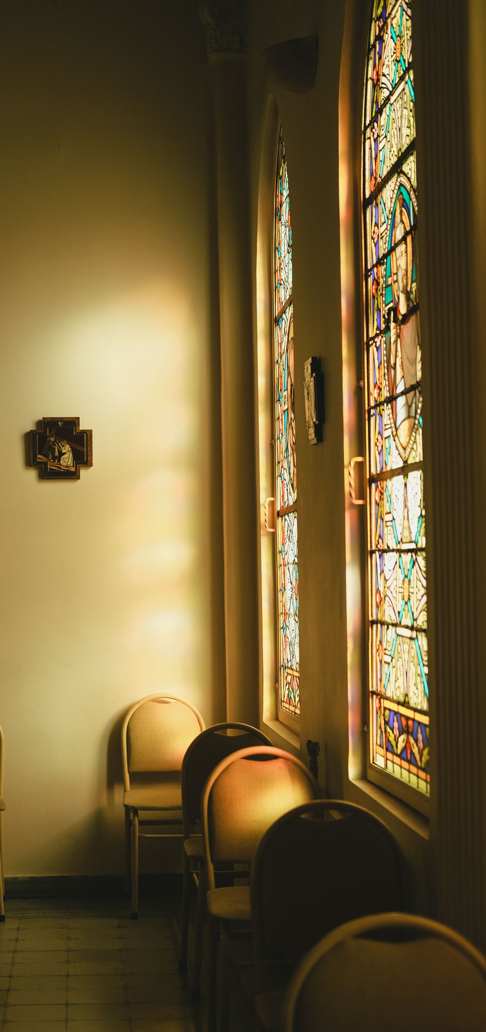Afternoon light through the stained-glass windows of a small chapel, with a devotional cross and stacking chairs.